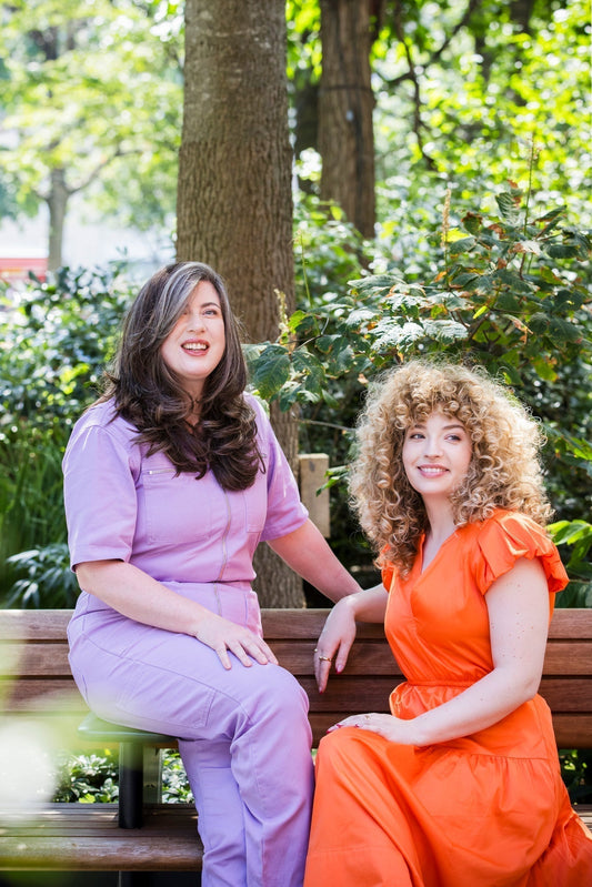 Two women in the 30s and 40s sitting on a bench smiling. One has smooth brunette and grey long hair with a purple jumpsuit and one has blonde curly hair is wearing an orange dress.